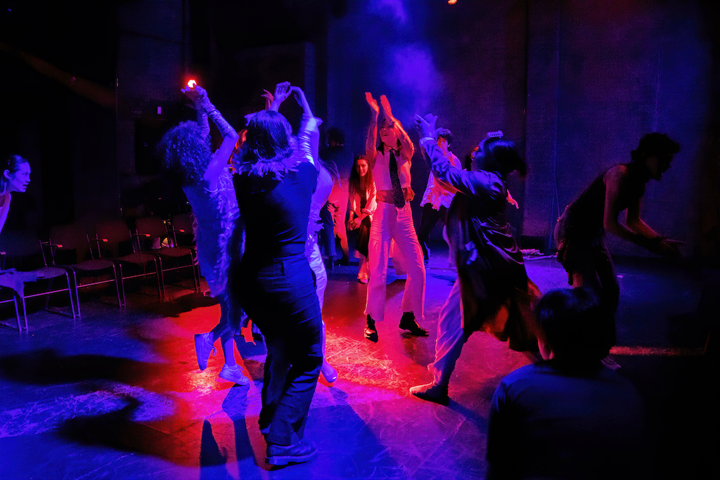 Actors lit with a red and purple spotlight stand with arms outstretched on stage in mid-performance